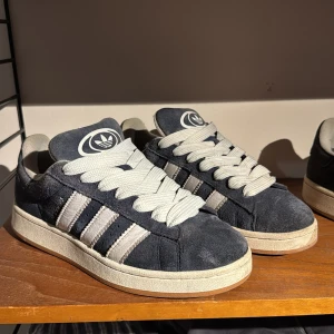 Adidas Campus sneakers i mörkblå mocka - Säljer ett par Adidas Campus sneakers i mörkblå mocka med klassiska vita ränder och breda vita skosnören. Skorna har en platt sula i gummi och rund tå. Adidas logga syns både på plösen och hälen. Perfekta för dig som gillar retrostil och streetwear. Kartong och kvitto finns