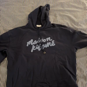 Mörkblå Maison Kitsuné hoodie M - Säljer en mörkblå hoodie från Maison Kitsuné i storlek M. Tröjan har huva med snörning och ett stort Maison Kitsuné-tryck i vitt på bröstet. Klassisk passform och mjukt material, perfekt för chill dagar eller när du vill ha en clean streetstyle.