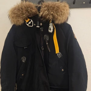 Svart vinterjacka Parajumpers med päls - Svart Parajumpers Anchorage vinterjacka med stor huva och fet pälsdetalj. Jackan har flera fickor, coola gula detaljer och patch på ärmen. Slim fit-modell med dragkedja och knappar framtill. Perfekt för kalla dagar och riktigt snygg streetstil. Står L men jag är själv medium å den sitter bra