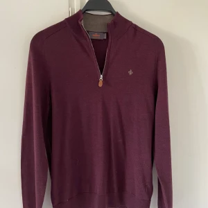 Vinröd merinoullströja från Morris - Säljer en vinröd långärmad tröja från Morris i 100% merinoull. Tröjan har half zip-dragkedja vid halsen, ribbade muddar och en broderad logga på bröstet. Perfekt för dig som gillar stilrena och bekväma plagg med snygga detaljer.