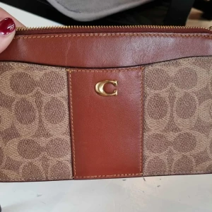 Brun plånbok från Coach i skinn - Snygg plånbok från Coach i brunt skinn och canvas med klassiskt monogrammönster. Guldiga detaljer, dragkedja upptill och flera kortfack på baksidan. Perfekt storlek för att ha med sig det viktigaste och stilren design med Coach-logga framtill.