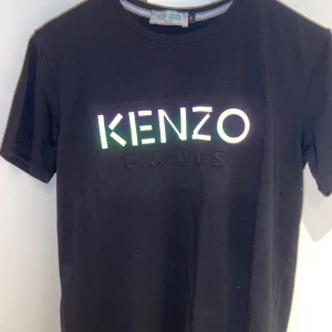 Svart Kenzo t-shirt med logga - Svart t-shirt från Kenzo med stor reflekterande logga på bröstet. Klassisk rund halsringning och korta ärmar. Tillverkad i mjuk bomull för en skön känsla. Perfekt för dig som gillar streetstyle och vill sticka ut med en ikonisk look.