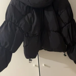 Svart pufferjacka med huva - Säljer en svart pufferjacka med huva och dragkedja framtill. Jackan har breda, puffiga ärmar och dragsko i nederkant för extra passform. Perfekt för kalla dagar och riktigt snygg till streetwear-stilen.