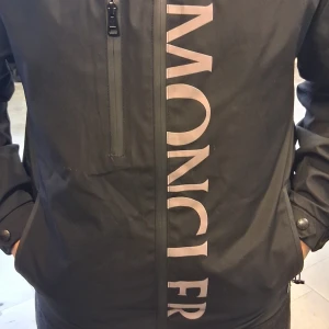 Svart vindjacka från Moncler med huva - Svart vindjacka från Moncler med stor logga framtill, huva och dragkedja. Jackan har en bröstficka med dragkedja, två sidofickor och Moncler-märke på ärmen. Materialet är lätt och smidigt, perfekt för blåsiga dagar.