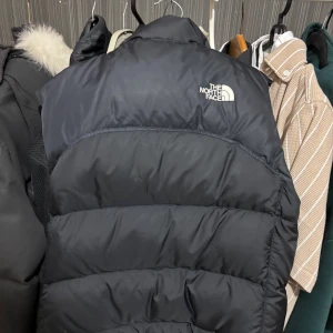 Svart dunväst från The North Face - Säljer en svart dunväst från The North Face med hög krage och broderad logga på bröstet. Västen har dragkedja framtill och två fickor med dragkedja. Klassisk pufferstil med 700-fill dun för extra värme. Perfekt för lager-på-lager under kyliga dagar.