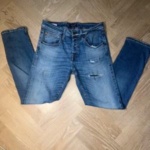 Blå jeans från Jack & Jones, 28/30 - Snygga blå jeans från Jack & Jones med slitna detaljer och klassisk femficksmodell. Jeansen har raka ben och normal passform, tillverkade i tåligt denim med coola slitningar framtill. Perfekta för en avslappnad streetstil.