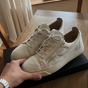 Vita sneakers med dragkedja och snörning - Säljer ett par vita sneakers från Giuseppe Zanotti med snygg glansig finish i skinn. Skorna har rund tå, snörning och coola dragkedjor på sidorna som ger en unik look. Perfekt för dig som gillar stilrena och trendiga sneakers med exklusiv känsla.