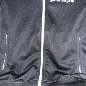 Svart track jacket från Palm Angels - Palm Angels svart track jacket med vita detaljer och logga på bröstet. Jackan har vita ränder längs ärmarna, hel dragkedja framtill och fickor med dragkedja. Tillverkad i polyester för en sportig vibe. Perfekt för dig som gillar streetwear och snygga detaljer.