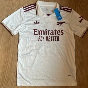 Arsenal Gabriel #6 vit Adidas tröja - Snygg Arsenal fotbollströja från Adidas i vit med vinröda detaljer och tryck. Kortärmad med klassiska Adidas-ränder på axlarna, broderat klubbmärke och Emirates Fly Better på bröstet. Gabriel och nummer 6 på ryggen. Skriv privat för mer frågor 