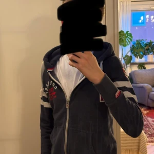 Blå Superdry hoodie med dragkedja - Säljer en blå hoodie från Superdry med vita ränder på ärmarna, vit snörning i huvan och röd/vit logga på bröstet. Tröjan har dragkedja framtill, känguruficka och är långärmad. Perfekt för dig som gillar sportig och avslappnad stil.