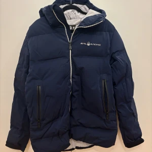 Marinblå pufferjacka från Sail Racing - Snygg marinblå pufferjacka från Sail Racing med huva och hel dragkedja. Jackan har två stora fickor med dragkedja framtill och logga på bröstet. Perfekt för kalla dagar med sin vadderade design och sportiga look.