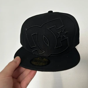 Svart DC Shoes keps 58,7cm - Säljer en svart keps från DC Shoes med stort broderat DC-logo framtill. Klassisk flatbrim-modell med guldfärgad klistermärke på skärmen. Kepsen är tillverkad i bomull och har en stilren, helsvart look.