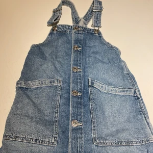 Jeansklänning med knappar från H&M - Säljer en ljusblå jeansklänning från H&M med stora fickor framtill och snygga metallknappar hela vägen ner. Klänningen har justerbara axelband och är tillverkad i bomull med lite stretch. Perfekt för en chill och avslappnad look.