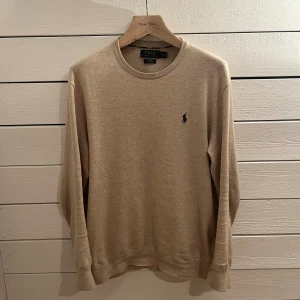 Finstickad tröja från Polo Ralph Lauren - Snygg beige tröja från Polo Ralph Lauren i slim fit med rund hals och diskret broderad logga på bröstet. Tillverkad i mjuk pima bomull, perfekt för dig som gillar stilrena och klassiska plagg med en modern touch.