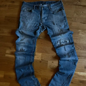 Distressed blå jeans från Armani Jeans - Säljer ett par blå jeans från Armani Jeans med slitna detaljer och trasiga partier på benen. Jeansen har klassisk femficksdesign, normal passform och är tillverkade i bomull. Perfekt för dig som gillar en edgy och avslappnad stil.