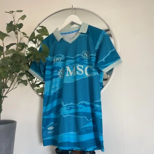 Napoli EA7 blå fotbollströja S - Napoli fotbollströja från EA7 i blått med ljusblå detaljer och mönster som påminner om vågor och fiskfjäll. Tröjan har korta ärmar, klassisk krage och tryckta loggor som MSC och Partenopei. Tillverkad i lätt och ventilerande material, perfekt för match eller träning.