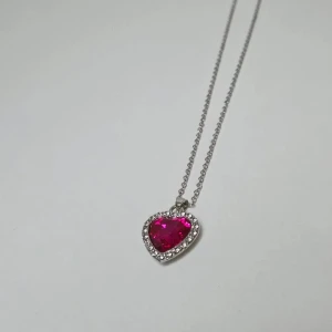 Halsband med hjärthänge och rosa sten 45 cm - Snyggt halsband i silverfärg med ett hjärtformat hänge. Hänget har en stor rosa glittrig sten i mitten, omgiven av små klara stenar som ger extra bling. Baksidan av hänget är texturerad i silver. Perfekt accessoar för dig som gillar statement-smycken.