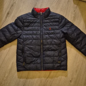Klassisk mörkblå och röd pufferjacka Polo Ralph Lauren - Snygg mörkblå pufferjacka från Polo Ralph Lauren med röd broderad logga på bröstet och röd insida den är vändbar. Jackan har dragkedja framtill, två fickor med dragkedja och quiltad design. Tillverkad i återvunnen nylon, perfekt för kyliga dagar. Ny pris 2395 tusen.