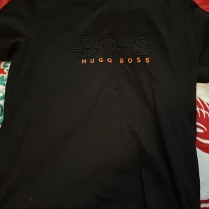 Svart t-shirt från Hugo Boss - Svart t-shirt från Hugo Boss med stor BOSS-logga i svart och texten HUGO BOSS i orange framtill. Klassisk passform och rund hals. Perfekt för dig som gillar stilrena och snygga märkesplagg.
