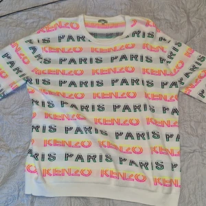Vit Kenzo tröja med neonränder - Cool vit tröja från Kenzo med färgglada neonränder och texten 'KENZO PARIS' i svart och neonfärger över hela tröjan. Rund hals och ribbade muddar. Perfekt statement-plagg för dig som gillar att sticka ut med färg och mönster.