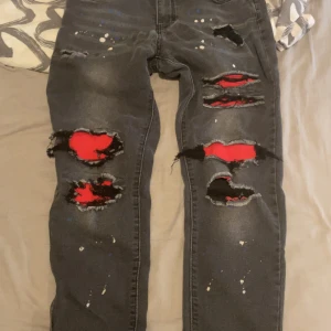 Svarta jeans med röda detaljer och slitningar XS - Unika svarta jeans med stora slitningar och röda tygdetaljer under hålen. Byxorna har färgstänk i vitt och blått för en extra edgy look. Modellen är rak och materialet är klassisk denim. Perfekta för dig som vill sticka ut med din stil. XS