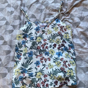 Blommig linnetopp från Angelology. Perfekt på sommaren - Supersöt linnetopp från Angelology med blommigt mönster i blått, gult och rött på vit botten. Tunt tyg med smala axelband och v-ringning. Perfekt för varma dagar och enkel att styla med jeans eller kjol.