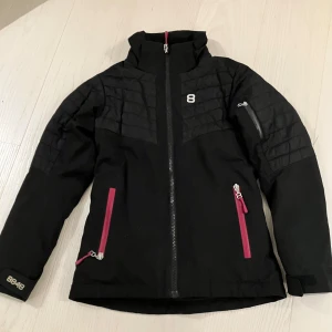 Svart vinterjacka skidjacka 8848 Altitude 140cm - Svart vinterjacka från 8848 Altitude med quiltade partier upptill och coola rosa dragkedjor. Jackan har hög krage, långa ärmar och praktiska fickor. Perfekt för kalla dagar och snygg streetstil samt till backen. 