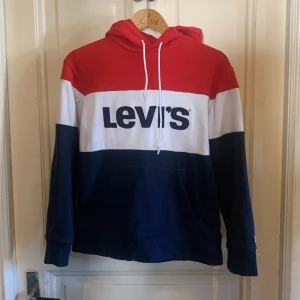 Levi's hoodie i rött, vitt och blått - Säljer en snygg hoodie från Levi's i storlek S. Tröjan har breda blockränder i rött, vitt och mörkblått, stor broderad Levi's-logga på bröstet och huva med vit snörning. Mjuk bomullskänsla och känguruficka framtill. Perfekt för chill dagar.