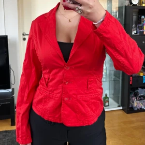 Röd kavajjacka från b.young - Snygg röd kavajjacka från b.young med klassisk krage, tre knappar framtill och fickdetaljer. Jackan har markerad midja och dekorativ knappdetalj bak. Perfekt för dig som vill sticka ut med färg och stil. Står ingen storlek men skulle säga S. Skriver måtten här. Axelbredd: 42cm. Längd: 60cm. Byst: 49cm.