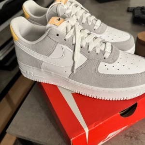 Nike Air Force 1 i vitt och grått mocka - Nike Air Force 1 sneakers i en clean vit och ljusgrå färgkombination med detaljer i gult på hälen. Ovandel i skinn och mocka, klassisk snörning och perforerad tå för extra ventilation. Snygg och tidlös siluett med platt sula och ikonisk Nike-logga på sidan.