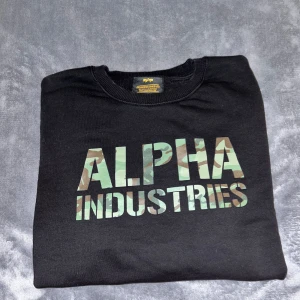 Svart Alpha Industries sweatshirt - Cool svart sweatshirt från Alpha Industries med kamouflagemönstrad logga på bröstet. Tröjan har lång ärm och en unik ficka med röd tag på ena ärmen. Tillverkad i mjuk bomull, perfekt för en streetwear-look.