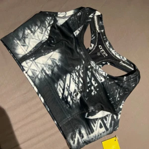 Svartvit sporttopp från OTP storlek s - Sportig svartvit topp från OTP med coolt abstrakt mönster. Modellen är ärmlös med brottarrygg och har en tight passform som passar perfekt till träning. Tillverkad i stretchigt syntetmaterial för maximal komfort och rörelsefrihet.