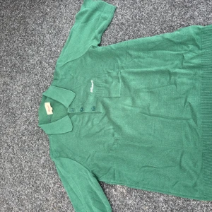 Grön pikétröja från Manors - Snygg grön pikétröja från Manors med klassisk krage, tre knappar och liten bröstficka. Tröjan har korta ärmar och är tillverkad i mjukt stickat material. Perfekt för dig som gillar en clean och sportig look.