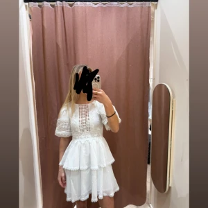 Vit spetsklänning med volanger - Superfin vit kortklänning med spetsdetaljer och volanger. Klänningen har korta ärmar, markerad midja och vackra broderade partier framtill och på ärmarna. Den dubbla volangkanten ger en extra söt och romantisk look. Perfekt för sommarens alla tillfällen eller studenten!