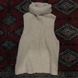 Beige stickad väst med polokrage - Mysig beige stickad väst med hög polokrage och ribbad struktur. Perfekt att slänga över en skjorta eller långärmad tröja för extra värme och stil. Enkel och clean design som passar till det mesta.