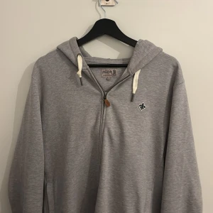 Grå hoodie från Morris med dragkedja - Säljer en stilren grå hoodie från Morris i storlek L. Tröjan har huva med vita snören, dragkedja framtill och en liten broderad logga på bröstet. Perfekt för en chill och avslappnad look.