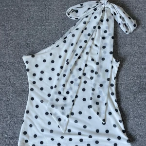 Prickig one shoulder topp  - Säljer en vit one shoulder topp från H&M med svarta prickar och stor knytrosett vid axeln. Superpopulär och slutsåld, endast använd en gång! (Man kan även ha bandet runt halsen) 