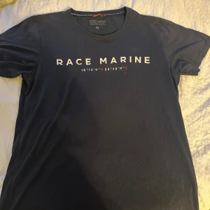 Mörkblå Race Marine t-shirt - Snygg mörkblå t-shirt från Race Marine med vit logga och koordinater tryckta på bröstet. Klassisk rund halsringning och korta ärmar. Perfekt för dig som gillar stilren och sportig design.