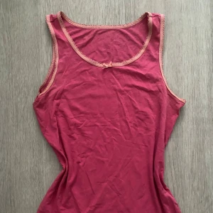 Rosa linnetopp med rosett och spetskant - Söt rosa linnetopp med ljusrosa spetskant runt halsringning och ärmhål. Dekorerad med en liten rosett framtill. Toppens material känns mjukt och stretchigt, perfekt för varma dagar. Enkel och feminin design som är lätt att matcha. xs/s