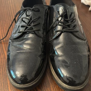 Svarta lackskor från Tamaris, stl 39 - Säljer ett par svarta lackskor från Tamaris i storlek 39. Skorna har snörning, rund tå och en platt sula. De är tillverkade i syntetmaterial med en glansig finish som ger en stilren look. Perfekta för dig som vill ha en klassisk och trendig sko.