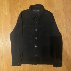 Svart overshirt från Donce De La Sense XS - Stilren svart overshirt från Donce De La Sense i storlek XS. Jackan har klassisk krage, två stora bröstfickor och knäppning framtill med svarta knappar. Materialet är mjukt och känns som ull, perfekt för lager-på-lager. Snygg broderad logga på ärmen.
