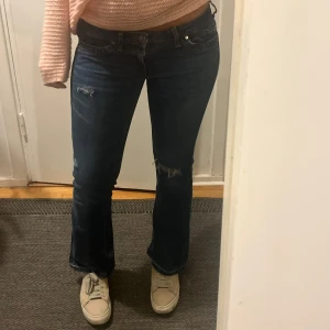 Lågmidjade jeans med hål - Säljer ett par mörkblå jeans med bootcut-modell och snygga slitningar på låret och knät. Jeansen har klassisk femficksdesign, låg midja och är tillverkade i ett stretchigt denimtyg som sitter skönt. Perfekta för dig som gillar en avslappnad men trendig look.