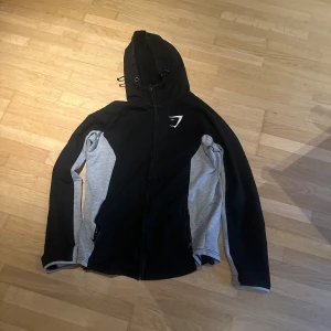 Svart och grå hoodiejacka från Gymshark - Svart och grå hoodiejacka från Gymshark med dragkedja framtill och två fickor. Jackan har lång ärm, huva med snörning och Gymshark-logga på bröstet. Tillverkad i mjuk bomullsmix, perfekt för träning eller chill.  Pris kan diskuteras