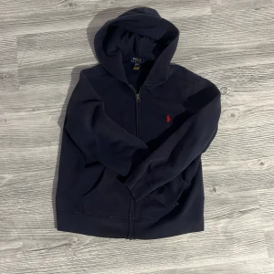 Mörkblå hoodie från Polo Ralph Lauren - Mörkblå hoodie från Polo Ralph Lauren med dragkedja och huva. Tröjan har ett rött broderat logomärke på bröstet, två fickor fram och är fodrad med mjuk fleece. Perfekt för kyliga dagar och enkel att matcha med jeans eller joggers.