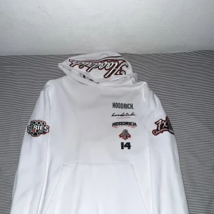Vit hoodie från Hoodrich med brodyr - Vit hoodie från Hoodrich med broderade detaljer och tryck på både fram- och baksida. Stora loggor, text och coola patches på ärmarna. Huva med broderad text och klassisk känguruficka framtill. Perfekt för dig som gillar streetwear vibes.
