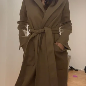 Lång coat med bälte  - Snygg beige kappa med bred krage och knytskärp i midjan. Lång modell med stora, praktiska fickor framtill och markerad söm i ryggen. Storlek 34/XS (endast använd en gång)