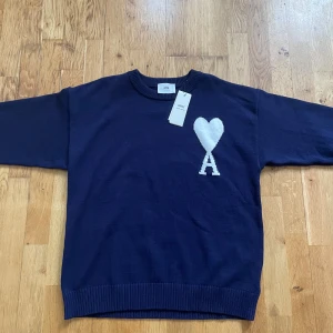 Mörkblå stickad tröja från Ami - Säljer en mörkblå stickad tröja från Ami med deras ikoniska vita hjärta och A-logga på bröstet. Tröjan har rund halsringning, ribbade muddar och är långärmad. Perfekt för dig som gillar stilrena och trendiga plagg.