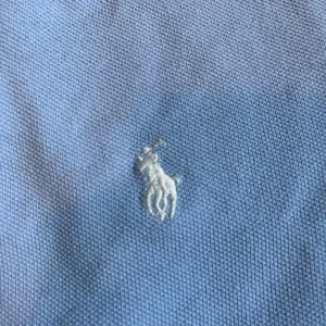Ljusblå pikétröja från Polo Ralph Lauren - Snygg ljusblå pikétröja från Polo Ralph Lauren med klassisk krage, två knappar och den ikoniska broderade polospelaren på bröstet. Tröjan är i bomull och har korta ärmar, perfekt för en clean och stilren look.