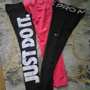 Nike leggings svart & rosa, Just Do It - Två par Nike leggings, ett svart med stor vit 'JUST DO IT.' text längs benet och ett par helsvarta med vit Nike-logga och bred resår med 'NIKE PRO' upptill. Det tredje paret är rosa med liten vit Nike-logga. Perfekta för träning eller chill.
