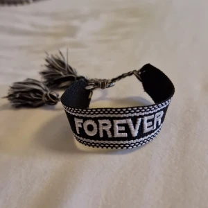 Svart och vit FOREVER armband - Snyggt vävt armband i svart och vitt med texten FOREVER broderad framtill. Armbandet har tvinnade band och tofsar i ändarna för en cool och personlig look. Perfekt accessoar för att lyfta din outfit med ett statement. Aldrig använd, inte riktigt min stil.